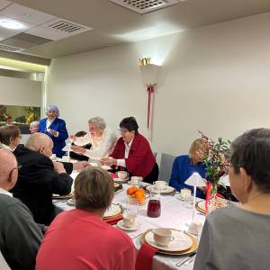 Wigilia w Eurohotelu. Moment łamania się opłatkiem. Seniorki i seniorzy siedzący po dwóch stronach stołu, wstają i składają sobie życzenia.