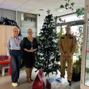 W holu Centrum Usług Społecznych przy ubranej choince od lewej stoją: pracowniczka Centrum, dyrektor Centrum i strażak. Przed nimi leżą prezenty.