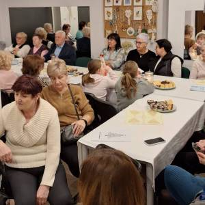 Zdjęcie przedstawia dużą grupę seniorów siedzących przy stołach nakrytych białymi obrusami, na których stoją talerze z wypiekami i zapalona świeca.