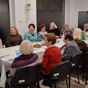 Zdjęcie przedstawia seniorki siedzące po dwóch stronach stołu nakrytego białym obrusem, na którym stoją talerze z ciastkami i ozdoba świąteczna.