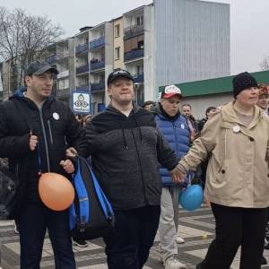 Na zdjęciu widać cztery osoby z niepełnosprawnościami, które idą po deptaku trzymając się za ręce. W tle widać sklep z blachy i blok mieszkalny.