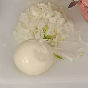 Zdjęcie przedstawia zbliżenie na obraną cebulę leżącą w całości na blacie, na małe kawałki już pokrojonej cebuli i dłonie trzymające nożyk i krojące kawałek warzywa.