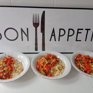 Na zdjęciu widać trzy białe głębokie talerze, w których jest makaron świderki, boczek i warzywa. Talerze stoją na tle tablicy z napisem Bon Apetit.