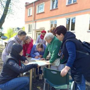 Grupa ośmiu seniorek przy jednym ze stanowisk gry terenowej. W tle widać budynek Dziennego Domu Senior Wigor i drzewo.