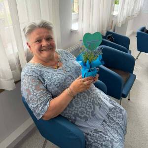 Zdjęcie przedstawia seniorkę siedzącą na fotelu, trzymającą w ręku niezapominajkę w doniczce.