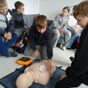 Dwóch chłopców klęczy na podłodze przy fantomie. Jeden z nich przylepia do fantoma elektrody od sprzętu AED, a drugi obserwuje akcję.