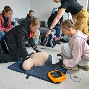 Gra miejska pod tytułem Ratujemy, bo umiemy. Na podłodze klęczą dwie dziewczynki. Jedna z nich układa fantoma. Druga trzyma w rękach elektrody AED.