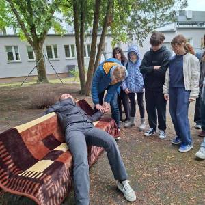 Ogród społeczny przy ulicy Poznańskiej. Grupa uczniów stoi przy ławce, na której leży aktorka udająca osobę nieprzytomną. Jeden z uczniów sprawdza jej puls.