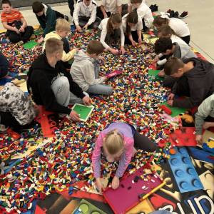 Kilkanaścioro uczniów i uczennic siedzi w wielkiej ilości różnokolorowych klocków Lego rozsypanych na podłodze i tworzy własne budowle.
