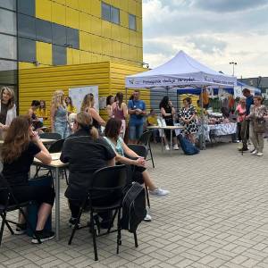 Plac przed szkołą. Na pierwszym planie widać trzy kobiety siedzące przy stole, dalej osoby stojące i przechodzące przy namiocie Hospicjum Palium.