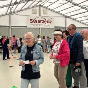 Na pierwszym planie uradowani seniorzy czekają w kolejce do konkurencji. W tle widać seniorów wykonujących slalom z kijem i piłeczką do hokeja.