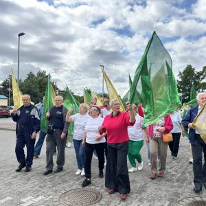Senioriada. Korowód uczestników z kolorowymi flagami idzie przez parking przed pływalnią. Na pierwszym planie widać dyrektor Centrum Usług Społecznych.