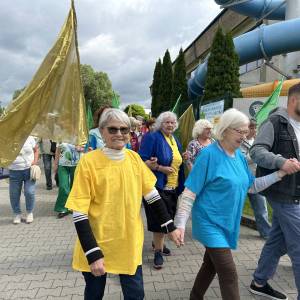 Senioriada. Korowód kolorowo ubranych seniorów niosących kolorowe flagi. W tle widać budynek pływalni Wodny Raj.