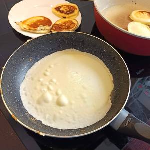 Zdjęcie przedstawia dużą patelnię, na której smaży się rozlane ciasto naleśnikowe. Na cieście widać charakterystyczne bąble powietrza.
