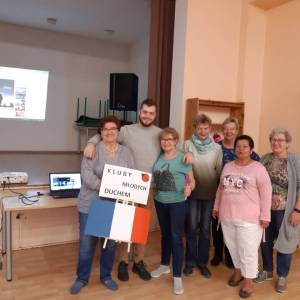 Wnętrze Centrum Aktywności Seniora. Wolontariusz europejski stoi w otoczeniu uśmiechniętych seniorek. Jedna z nich trzyma flagę Francji i transparent.