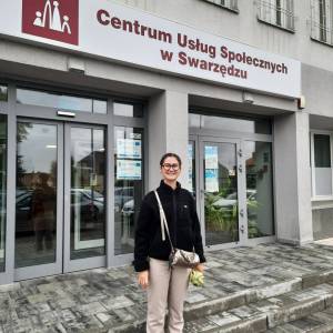 Uśmiechnięta wolontariuszka z Francji stoi przed głównym wejściem do Centrum Usług Społecznych. Mokry chodnik wskazuje, że przed chwilą padał deszcz.