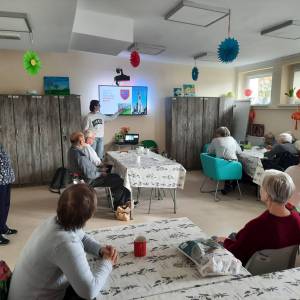 Główna sala w Dziennym Domu Pomocy. Seniorzy siedzą przy stolikach i słuchają wolontariuszki. Francuzka pokazuje coś na ekranie multimedialnym. 