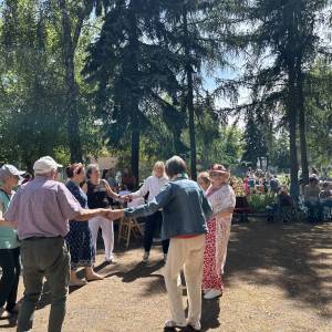 Ogród społeczny. W cieniu wysokich modrzewi, świerka i brzozy seniorzy tańczą w kółeczku.