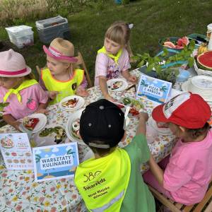 Kolorowe stanowisko warsztatów kulinarnych. Grupa przedszkolaków wykonuje pizzę z arbuza.