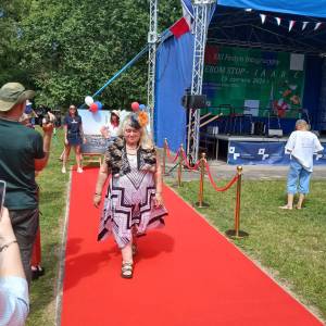 Festyn w Lisówkach. Uczestniczka Domu Senior Wigor w eleganckiej kreacji z fascynatorem na głowie prezentuje swoją osobę idąc po czerwonym dywanie.