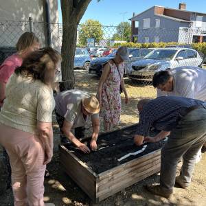 Seniorzy razem z prelegentem nawożą ziemię w jednej z drewnianych skrzyń.