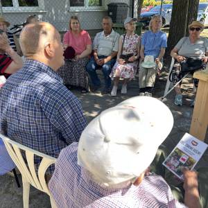 Seniorzy i seniorki siedzący w cieniu na krzesełkach słuchają prelekcji dotyczącej uprawy i nawożenia roślin.