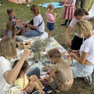 Śniadanie na trawie. Widoczny koc, na którym ułożono wiele różnych ozdób potrzebnych do zrobienia wianka. Dookoła koca zgromadziły się mamy z dziećmi.