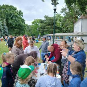 Śniadanie na trawie. Park przed Ośrodkiem Kultury. Na pierwszym planie dzieci i rodzice przy stanowisku Laboraków. W tle wiata rowerowa, ludzie, drzewa.