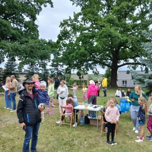 Park przed Ośrodkiem Kultury. Około trzydziestu osób stoi w różnych miejscach dookoła stanowiska malowania buzi Maxi Fun. W tle dwa rozłożyste dęby.