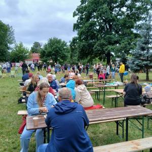 Park przed Ośrodkiem Kultury. Na drewnianych ławkach przy drewnianych stołach siedzą uczestnicy pikniku. W tle zieleń drzew i pozostali piknikowicze.