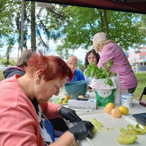 Zbliżenie na kobietę krojącą owoce. Na dalszym planie stół, na nim pomarańcze, cytryny, krzew bazylii, jogurt, rękawiczki, miska i trzy osoby.