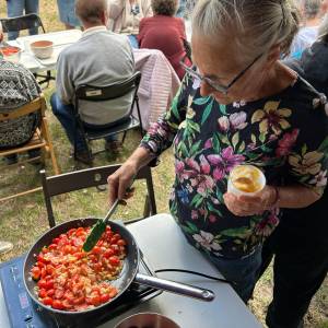 Warsztaty kulinarne. Jedna z seniorek szpatułką silikonową miesza pomidory z cebulką smażące się na patelni. W lewej ręce trzyma plastikowy kubek z koktajlem owocowym.