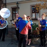 Zespół muzyczny w mundurach gra na instrumentach dętych na świeżym powietrzu, przed budynkiem w Swarzędzu. Na pierwszym planie widoczny jest dyrygent, a w tle baner z napisem 