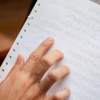 Zdjęcie przedstawia dłoń dotykającą kartki z tekstem zapisanym w alfabecie Braille’a. Palce przesuwają się po wypukłych punktach, które służą do czytania przez osoby niewidome i niedowidzące. Papier posiada perforowane brzegi, co sugeruje, że został wydrukowany na specjalnej drukarce Braille’a. Tło jest lekko rozmyte, a oświetlenie naturalne.