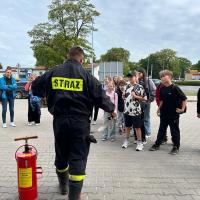 Zdjęcie przedstawia punkt startowy gry miejskiej pod tytułem Ratujemy bo umiemy. Na pierwszym planie widać strażaka tłumaczącego grupie uczniów konkurencję, do której za chwilę przystąpią. Obok strażaka stoi czerwona hydronetka wodna z żółtą etykietą.