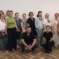 Zdjęcie wykonane w Centrum Międzypokoleniowym Pogodna. W sali na tle ściany stoi trzynaście przedstawicielek kół gospodyń wiejskich, kuca jeden mężczyzna również członek jednego z kół oraz organizatorka społeczności lokalnej.