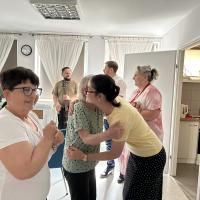 Wnętrze Dziennego Domu Pomocy. Pożegnanie Żuljet Żama wolontariuszki Europejskiego Korpusu Solidarności. Wolontariuszka przytula się do seniorki składającej jej życzenia. Druga seniorka czeka w kolejce do Żuljet. W tle widać troje pracowników.