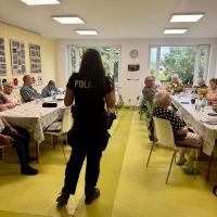 Zdjęcie przedstawia grupę seniorów siedzących przy stolikach zwróconych do policjantki, która stoi na środku pomieszczenia.