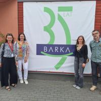 Czworo uczestników spotkania stoi na tle banera z napisem Barka 35 lat. Baner wisi na ścianie budynku, który jest siedzibą Fundacji Barka w Poznaniu.