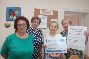 Wnętrze świetlicy wiejskiej w Paczkowie. Cztery seniorki pozują do zdjęcia trzymając plakaty projektowe.
