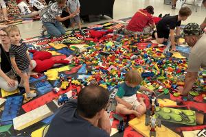 Piknik Trzech Pokoleń. Wnętrze namiotu przed sceną nad jeziorem. Na pierwszym planie widać rodziców z dziećmi bawiących się klockami lego, w tle widać kolejne stanowisko z klockami lego i sztalugi do malowania intuicyjnego.