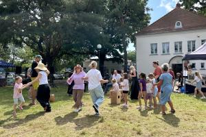 Zdjęcie wykonano na trawie przy budynku Ośrodka Kultury w Swarzędzu. Na pierwszym planie widoczne radośnie tańczące kobiety z dziećmi. W tle orkiestra flażoletowa, namioty z przekąskami i wysokie drzewa.