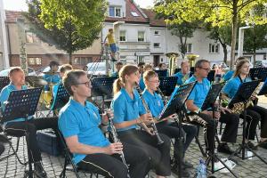 Kilkanaścioro muzyków ze Swarzędzkiej Orkiestry Dętej koncertują na Placu Niezłomnych w otoczeniu liściastych drzew i kamiennic. Na pierwszym planie muzycy grający na fletach prostych.