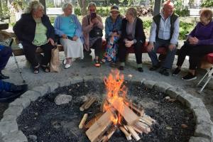 Na zdjęciu widać ognisko rozpalone w specjalnym palenisku pod wiatą. Przy palenisku złożone są porąbane klocki drzewa. Dookoła ogniska siedzą seniorki i seniorzy. W tle widać zielone drzewa, a za nimi wysoki jasny budynek z wieloma oknami.