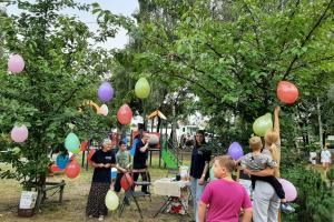 Zdjęcie przedstawia park rekreacyjny w Kobylnicy. Widać osoby zawieszające na drzewach balony różnych kolorów. Pod drzewami ustawione są krzesła i stoliki. W tle widać plac zabaw dla dzieci.