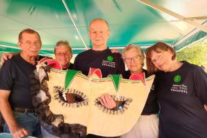 Zdjęcie przedstawia pięcioro uczestników festynu integracyjnego w Lisówkach - seniorki i seniorów z Dziennego Domu Senior Wigor trzymających przed sobą dużą ozdobną maskę karnawałową. Seniorzy mają na sobie granatowe tiszerty z logo placówki.