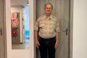 Zdjęcie przedstawia uśmiechniętego pana Jana stojącego na korytarzu Dziennego Domu Senior Wigor w Kobylnicy. Mężczyzna stoi przed drzwiami, nad którymi wisi jego dzieło - deska z wypalonymi motywami florystycznymi i hasłem Witaj Seniorze w Wigorze.