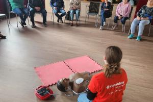 Wnętrze głównej sali w Centrum Aktywności Seniora. Na podłodze klęczy ratowniczka, która demonstruje seniorom użycie sprzętu do reanimacji. Przed nią na krzesłach w półokręgu siedzą seniorzy. Za nimi widać scenę, na której leży mnóstwo balonów.