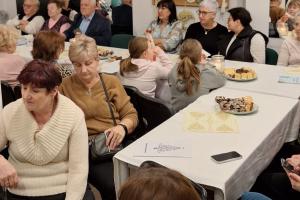 Zdjęcie przedstawia dużą grupę seniorów siedzących przy stołach nakrytych białymi obrusami, na których stoją talerze z wypiekami i zapalona świeca.