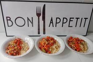 Na zdjęciu widać trzy białe głębokie talerze, w których jest makaron świderki, boczek i warzywa. Talerze stoją na tle tablicy z napisem Bon Apetit.